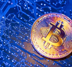 A golden Bitcoin coin resting on a blue illuminated circuit board with visible electronic pathways and solder points.