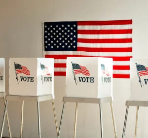 Voting booths labeled “VOTE” lined up indoors with a U.S. flag hanging on the wall behind them.