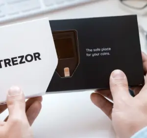 Hands holding an open Trezor hardware wallet box with the device visible on a desk.