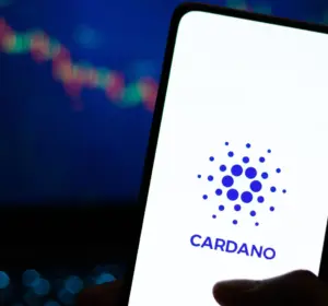 Hand holding a smartphone displaying the Cardano logo with a crypto price chart in the background