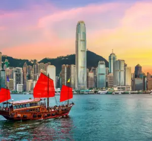 Traditional red-sailed junk boat crossing Victoria Harbour before Hong Kong’s skyline at sunset.