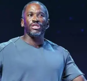 Man in a gray t-shirt speaking on stage while holding a presentation clicker, with a dark starry backdrop