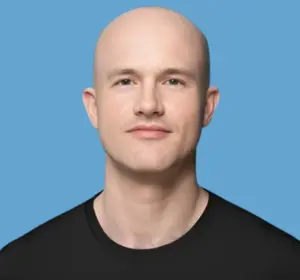 Coinbase CEO Brian Armstrong in a black t-shirt smiling slightly against a solid blue background