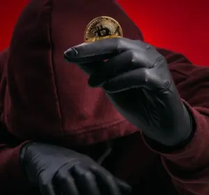 Hooded figure wearing black gloves holding a gold Bitcoin token against a red background