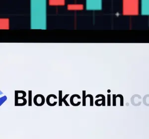 Blockchain.com logo displayed on a screen with red and green cryptocurrency trading charts in the background.