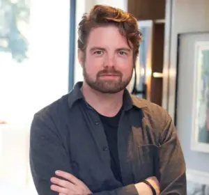 Man with wavy auburn hair and a trimmed beard stands with arms crossed in a bright, modern office, wearing a dark gray button-up shirt over a black t-shirt.