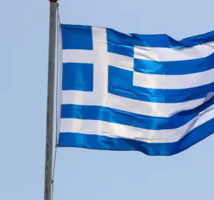 Greek national flag waving on a flagpole against a clear pale blue sky.