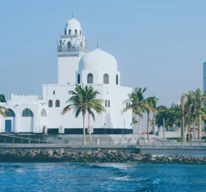 White waterfront mosque surrounded by palm trees beside the blue sea under a clear sky.